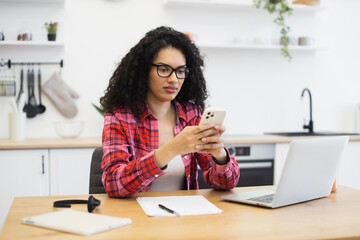 African woman wearing plaid shirt sitting at table using smartphone and laptop device multitasking in kitchen setting