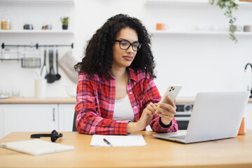 Young African woman wearing glasses in kitchen working remotely using smartphone and laptop,...