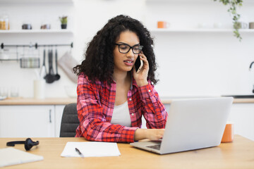 Young African woman in plaid shirt sitting at desk using laptop speaking on phone in home office....