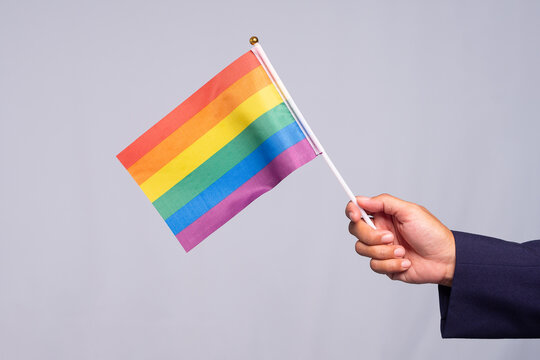 Hand holding a small rainbow pride flag against a neutral background.