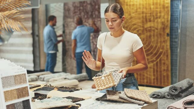 While visiting store with household and interior goods, girl examines examples of carpet products, chooses carpet material. She inspect small pieces of carpet, make decision about purchase. 