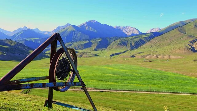 An old motorcycle wheel mounted on a metal triangular frame standing on a grassy mountain slope under the bright summer sun, with rolling green hills and distant mountains in the background.