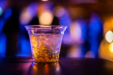 A plastic cup filled with whiskey and ice sits on a bar table, illuminated by vibrant bokeh lights in a nightclub atmosphere