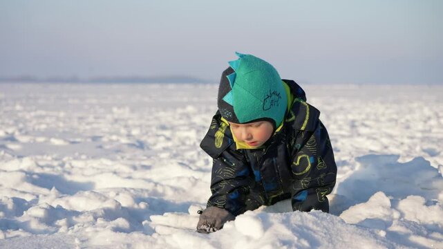 Young boy, bundled in warm winter clothes and a playful dinosaur hat, crawls through the snowdrifts on a vast frozen lake, his curiosity piqued by the icy wonderland surrounding him