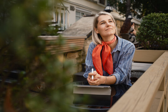A thoughtful woman sits outdoors at a cafe, wearing a red scarf and striped shirt. She appears relaxed and contemplative, enjoying a peaceful moment in the open air.
