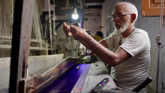 Old experienced Indian artisan hand weaving a Banarasi silk saree on a traditional wooden handloom. Adding thread to the weft shuttle