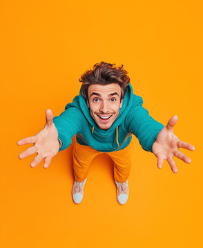  Photograph of a happy young man in a turquoise hoodie and yellow pants reaching out towards the camera, taken from a top-down perspective, against an orange background with stud