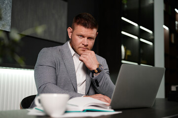 Obraz premium Confident businessman in creative office. Man in a suit with a serious expression looks into the camera lens with his fist under his chin. Manager works in an office at a desk.