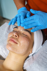 Beautician wearing blue gloves applying cleansing foam on young woman's face, preparing for...