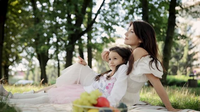 A mother and her young daughter enjoy a sunny day in the park, relaxing on the grass under trees. The warm atmosphere makes for a perfect family bonding moment in nature