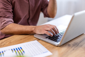 A man working on a laptop while analyzing data from a printed chart on a wooden desk. Home office concept, data analysis, productivity, and remote work.