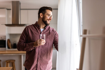 A man in casual wear drinks water in a modern kitchen, taking a moment to recharge. He stays...