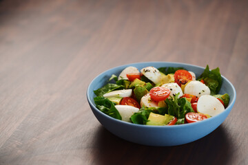 Salad with avocado, cherry tomatoes, romaine and mozzarella in blue bowl on walnut table with copy space