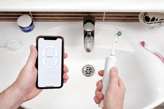 A man holds a smartphone and an electric toothbrush against bathroom sink.