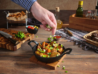 A man's hand sprinkles chopped green onions over cooked country-style potatoes in cast iron skillet...