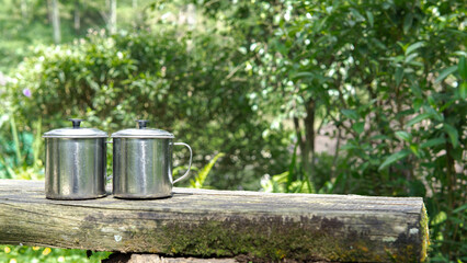 A pair of closed stainless mugs rest firmly on a mossy wooden table, blending harmoniously with the serene natural surroundings