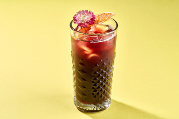 A vibrant cocktail with a flower garnish, served in a tall glass with ice on a bright yellow background.