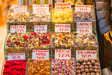 A vibrant selection of loose herbal teas at a Turkish spice market, displayed in glass containers with handwritten labels. The assortment includes pomegranate, rose, and energy tea.