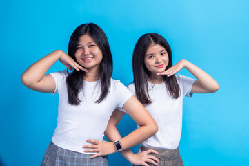 Two Asian teen girls smiling while posing with hands under their chins, standing in front of a...