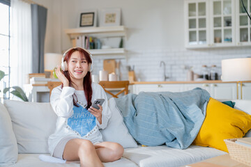 Asian stylish young woman is sitting on a sofa and listening to music with a wireless bluetooth...