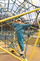 Happy child in a turquoise suit climbing on an alpine net on a playground on a spring day. Active outdoor games. Playground, sports ground for children.
