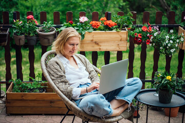 Young adult Caucasian blonde woman sitting on cottage terrace using laptop, online work in the countryside concept.  Intelligent attractive female successful freelancer does business in Her own home