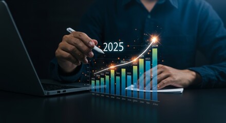 Hand with pen over 2025 bar graph, laptop.