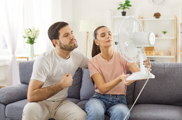 Unhappy young couple badly suffer with heat, using fan air for cooling or ventilation without...