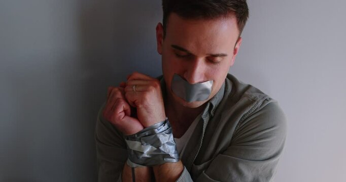 Portrait of young Caucasian male unconscious hostage tired with adhesive tape sitting on gray wall background. Stressed scared man victim loosed consciousness. Crime and kidnapping concept. 4k video.