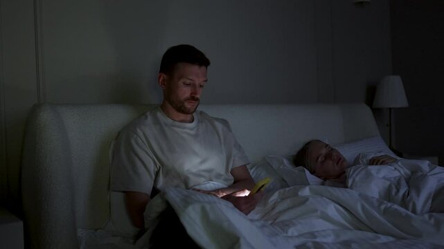 Cheating husband scrolls phone, chats on social networks, messaging in bed at night. Wife sleeps while man betrays absorbed in smartphone, web feed and digital addiction, irregular work schedule