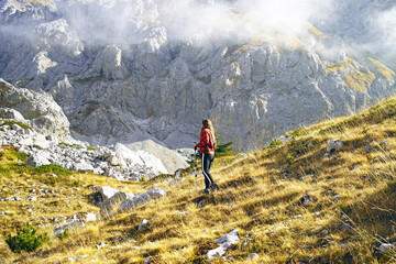 Exploring beautiful places in Montenegro: a female tourist during a walk in the Komovi Nature Park. A young woman with hiking poles goes down a slope against a backdrop of a mountain and clouds.