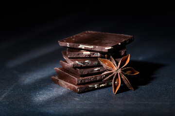 Dark chocolate chunks layering atop each other, complemented by star anise pods, resting on rustic dark surface highlighting rich culinary textures
