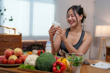 Asian sportswoman holding glass of milk smiling surrounded by healthy food in her apartment