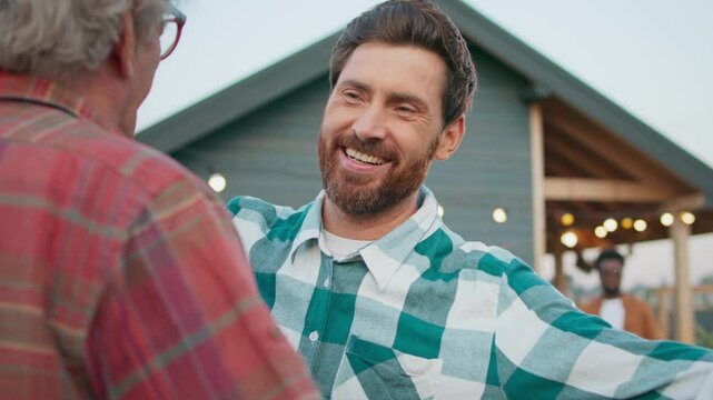Camera focus on Caucasian man in checkered shirt coming up to mature father. Men hugging each other while smiling. Family reunion. Son visiting dad in village. Lights shining on house in background.
