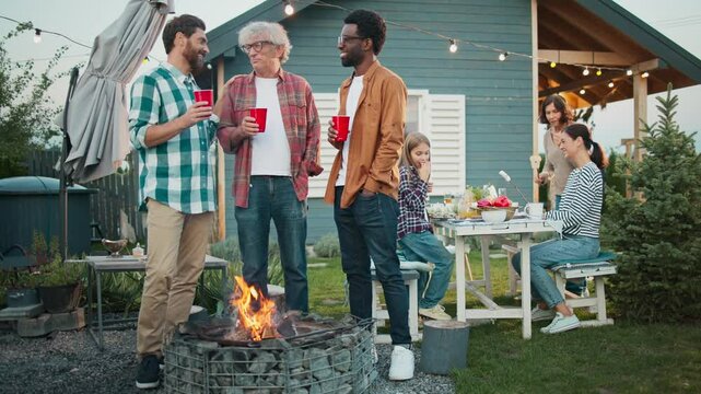 Cozy family gathering outside of house in countryside. Men standing in front of campfire while enjoying refreshing drink from red plastic cups. Women talking in background at table.