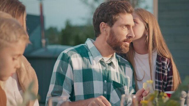 Warm moment of family enjoying food in fresh air. Bearded man sitting and preparing eating. Daughter whispering something into fathers ear. Dad smiling with joy while looking at beloved child.