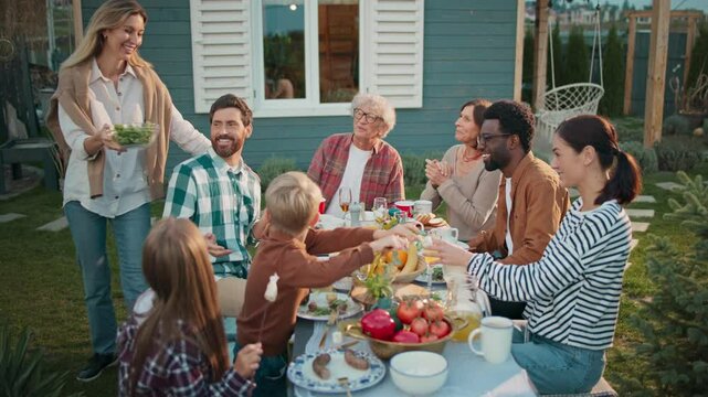 People gathering together at table. Woman bringing dish and placing to other plates. Family thanking woman about tasty food. Female blushing. Covering face with both hands. Picknick.
