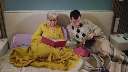 Elderly woman and young man reading books together while sitting on a bed in a cozy home setting.