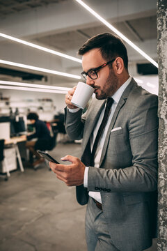 Positive middle aged businessman with beard in suit with cup of coffee using smartphone in office interior, free space. Break of work, video call, app for business, finance and gadget.