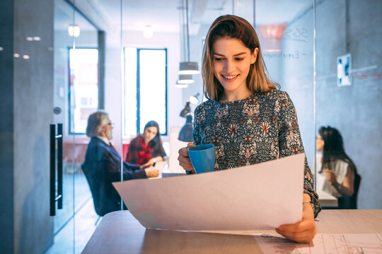 Portrait of happy beautiful young businesswoman looking at project and drinking coffee in front of her colleagues in the office.