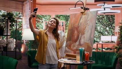 In modern atelier, artist filming rustic artwork painting with smartphone, social media branding. Recording and taking pictures of colorful canvas with watercolor piece, self promotion. Camera B.