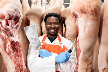 Portrait of professional butcher holding sharp cleaver and knife by raw meat hanging in...
