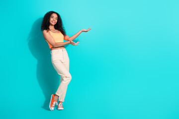 Smiling young woman in casual summer outfit presents with hands on a bright turquoise background