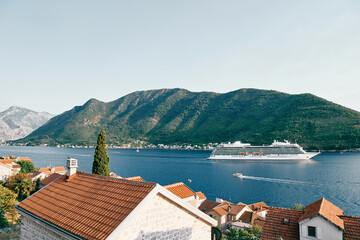 View over the red roofs of houses to a large white cruise ship sailing on the sea along a mountain ridge