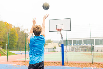 Back view of a man wearing sports clothes shooting free throw at basketball court. Unrecognizable young athlete playing basketball outside. Healthy lifestyle and outdoor sport concepts.