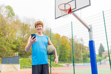 Smiling basketball player holding ball and using smartphone on court