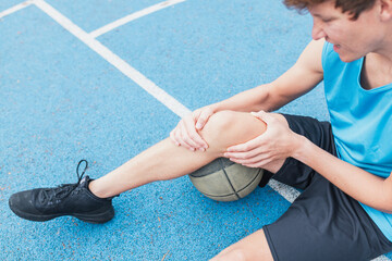 Man with basketball knee injury or pain sitting on the floor of basketball court. Man holding his...