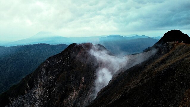 17.02.2025, Indonesia, North Sumatra. A group of hikers stands atop Sibayak volcano, surrounded by rugged volcanic rock. Steam rises from the crater. In the background city, mounitains and Sinabung.