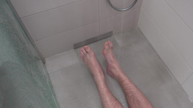 A man in the shower under a stream of water.
View of the legs of a man lying in the shower under running water. A man slipped on the floor in the shower. A man is lying on the floor in the shower.