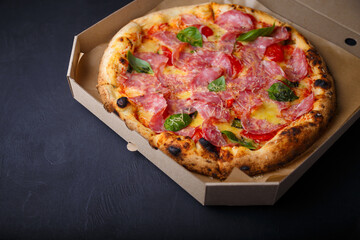Pizza with cheese, ham, green basil leaves and tomatoes in paper box on black background. Food product advertising banner, copy space